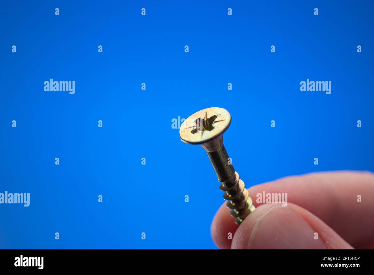 Petite vis filetée en métal maintenue entre les doigts par une main mâle caucasienne. Gros plan studio macro, isolé sur fond bleu Banque D'Images