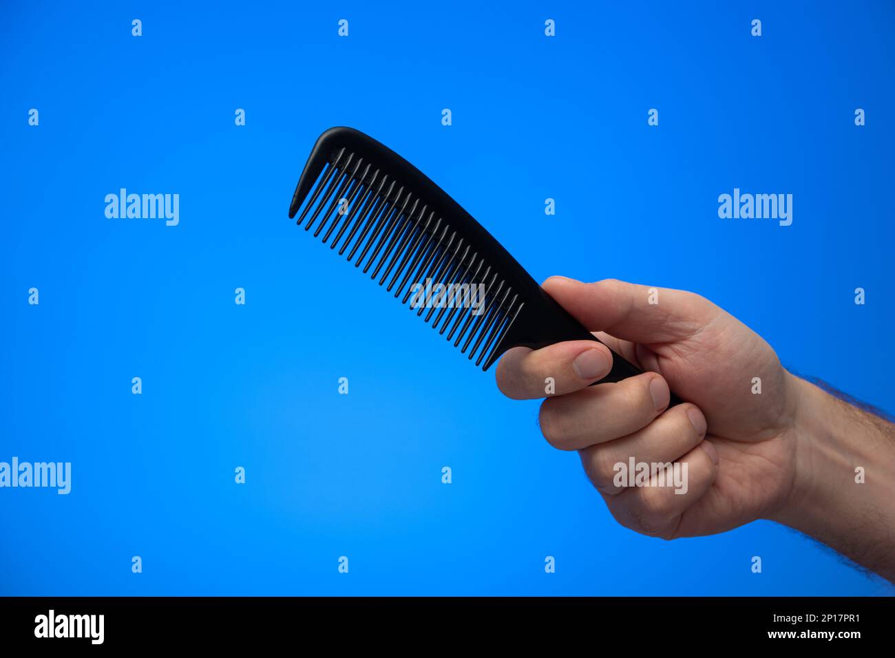 Main mâle caucasienne tenant un peigne à cheveux en plastique noir isolé sur fond bleu prise en studio. Banque D'Images