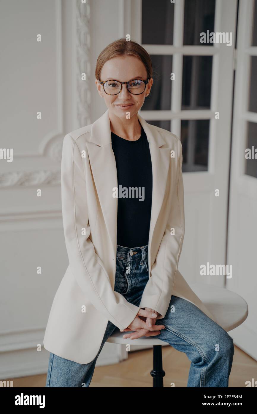 Sympathique femme d'affaires souriante dans des lunettes regardant l'appareil photo, assis sur une petite table ronde. Femme confiante portant des vêtements décontractés élégants, Banque D'Images