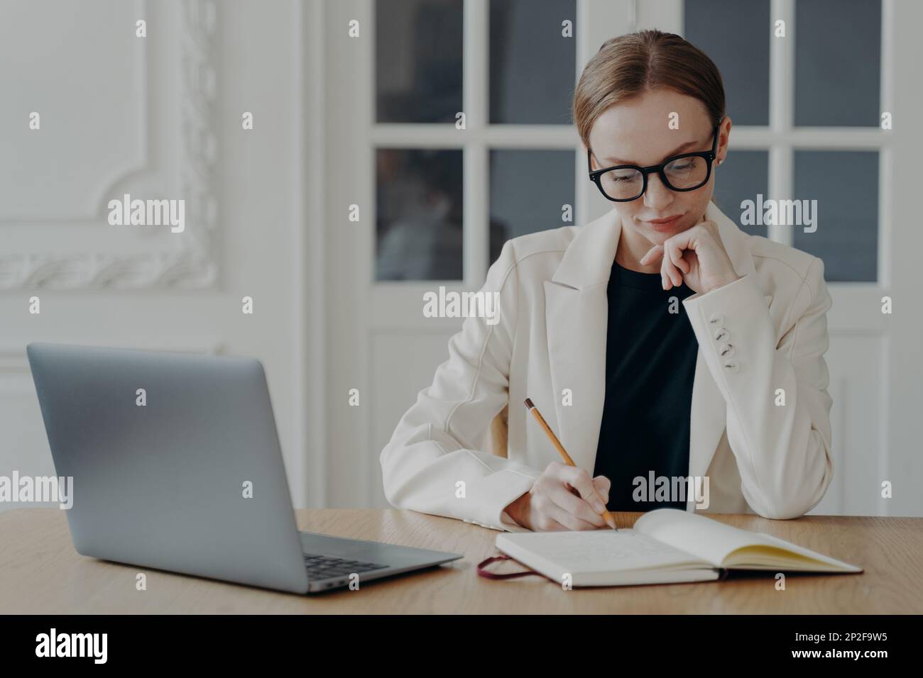 Femme d'affaires concentrée dans des lunettes écrivant dans un ordinateur portable, assis au bureau avec ordinateur portable. Femme tenant un crayon, prenant des notes, planifiant le calendrier d'entrée Banque D'Images