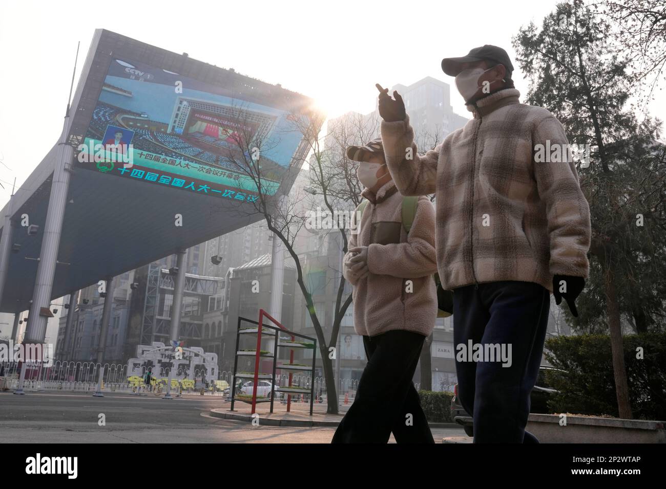 People wearing face masks walk past a large video screen showing the opening session of China's National People's Congress in Beijing, Sunday, March 5, 2023. (AP Photo/Andy Wong) Banque D'Images