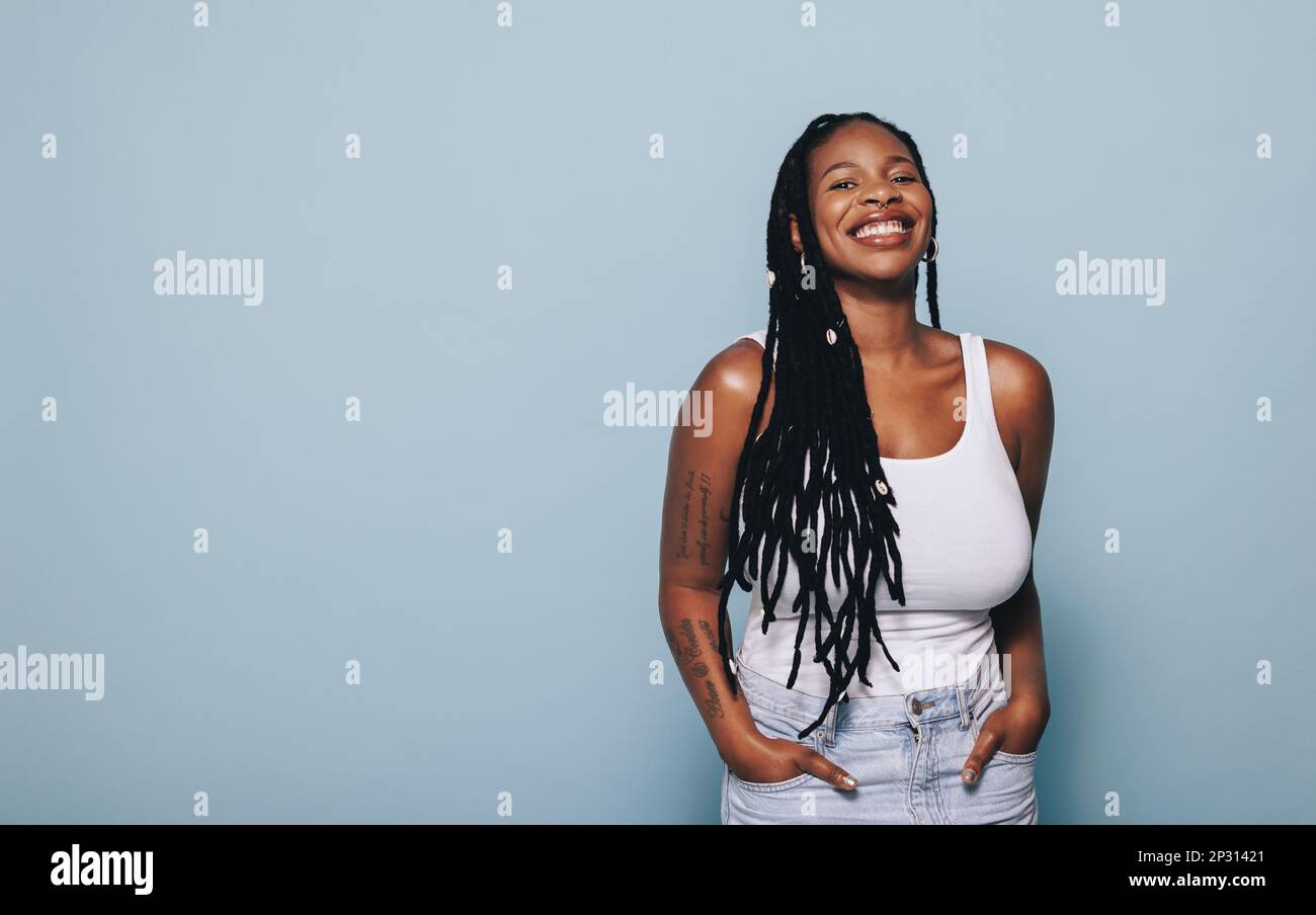 Jeune femme souriant à la caméra tout en se tenant debout sur un fond bleu. Jeune femme noire se sentant confiante dans son style. Banque D'Images