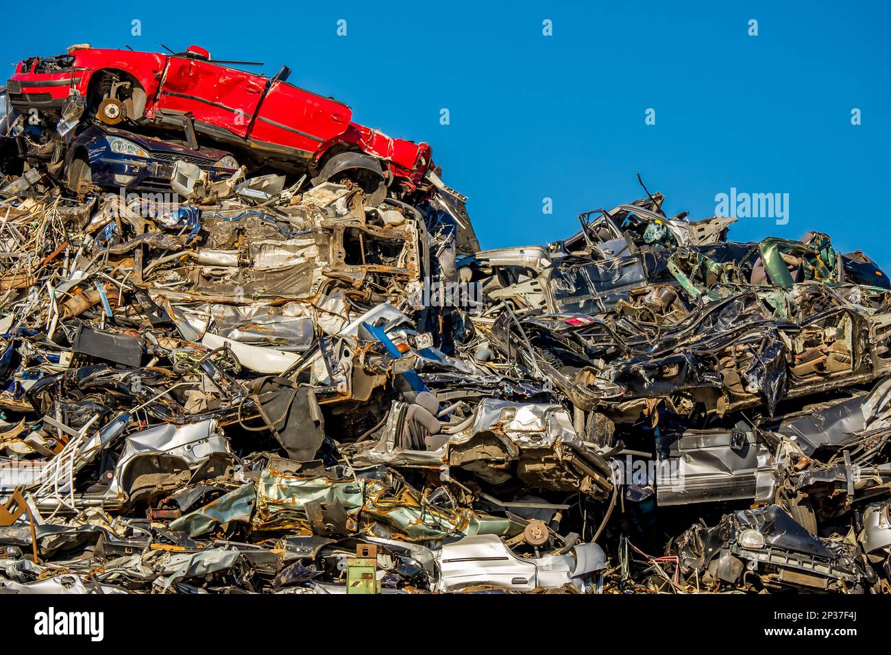 Une voiture rouge se trouve au-dessus d'une pile chaotique de voitures comprimées et écrasées dans un chantier naval, reflétant l'importance du recyclage des voitures et de la gestion des déchets. Banque D'Images