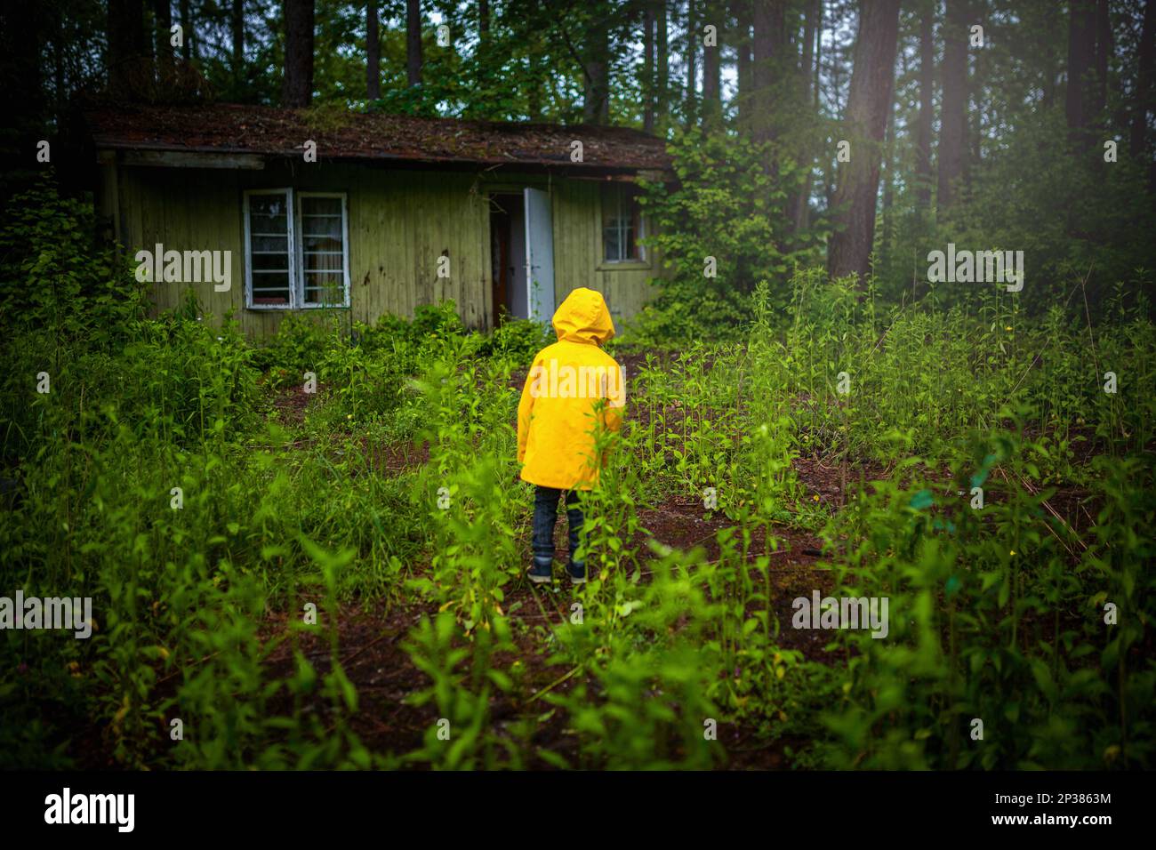 Un enfant dans un manteau jaune debout dans les bois en face d'une ancienne maison en bois. Un garçon de six ans Banque D'Images