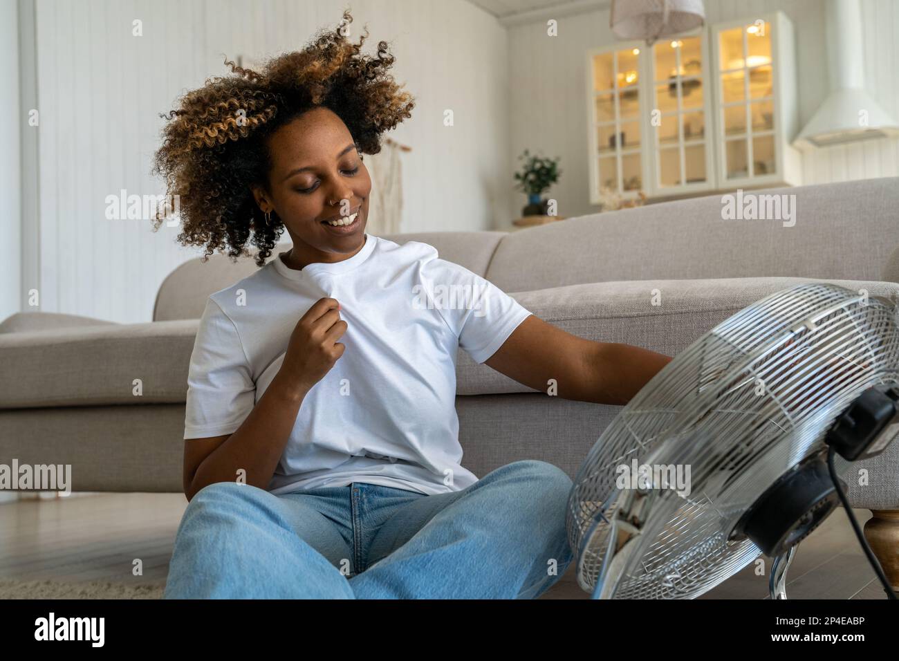 Heureuse joyeuse femme noire s'asseoir sur le sol refroidit maintient le ventilateur règle le flux d'air à la maison en journée de chaleur Banque D'Images