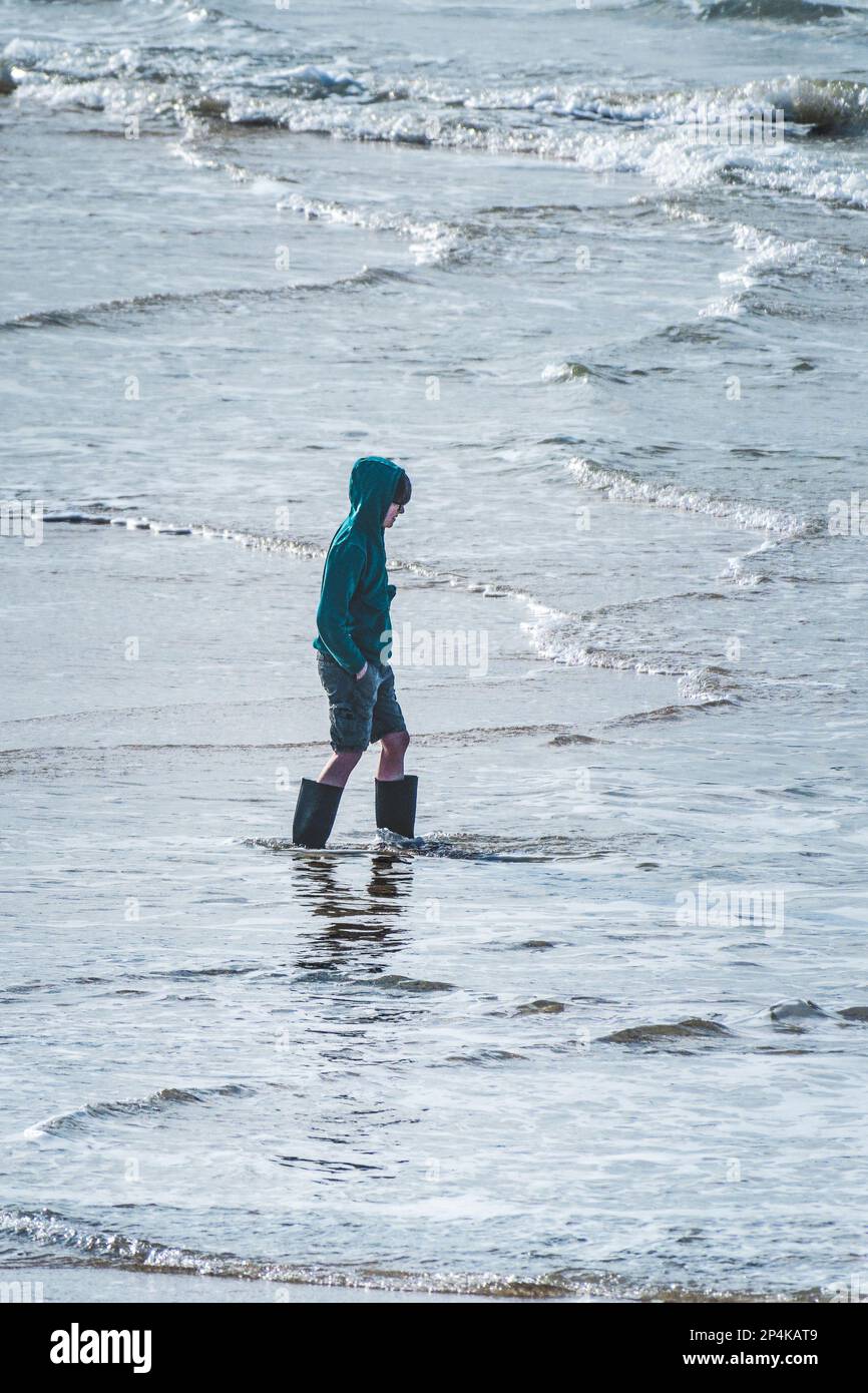 Un jeune garçon portant des bottes Wellington et barbotant dans la mer à Fistral, à Newquay, en Cornouailles, au Royaume-Uni. Banque D'Images