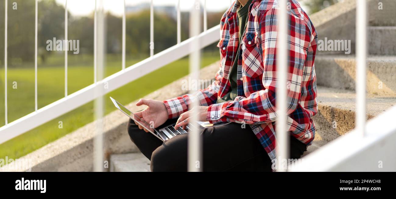 Personne d'apparence non binaire assise sur les marches à l'extérieur dans le parc et travaillant sur un ordinateur portable. Banque D'Images