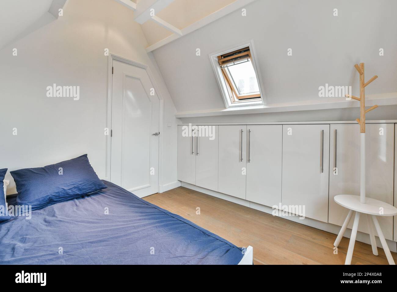 une chambre avec des armoires blanches et des draps bleus sur le lit en face de la fenêtre, il y a une petite table à côté Banque D'Images