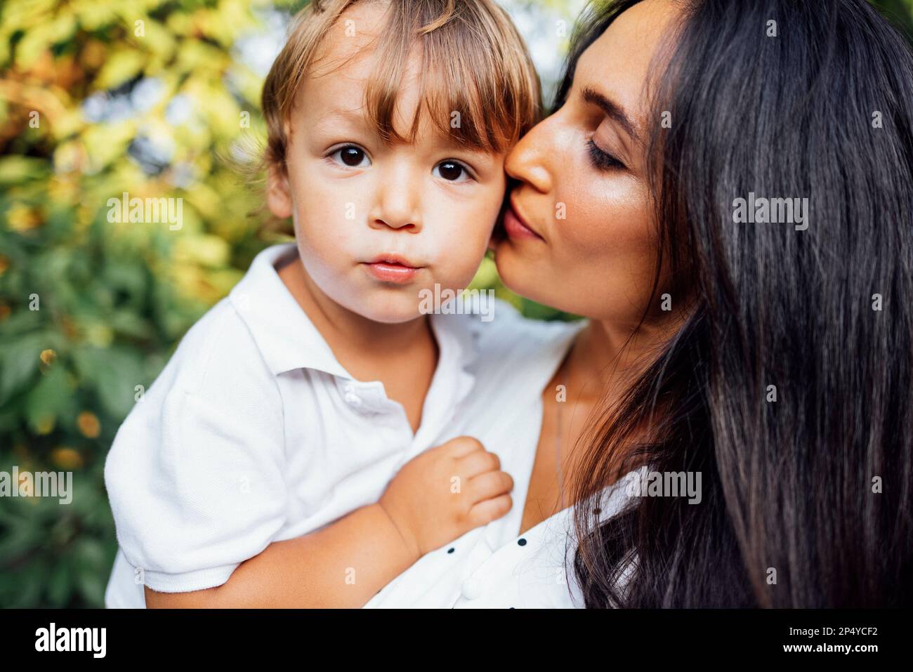 Joli portrait d'une belle mère embrassant son petit fils. Gros plan d'une jeune brune visage de femme touchant à son enfant mignon. Feuilles vertes dans le Banque D'Images