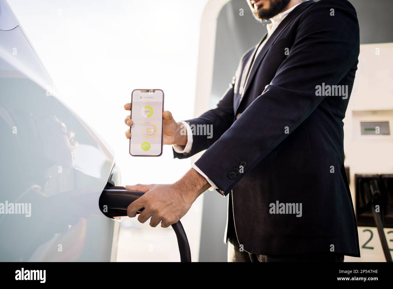 Récolte de l'homme en costume noir en branchant le cordon d'alimentation et en montrant l'écran mobile avec application de l'état de charge de la batterie de voiture électrique. Concept de technologie et d'innovation. Banque D'Images