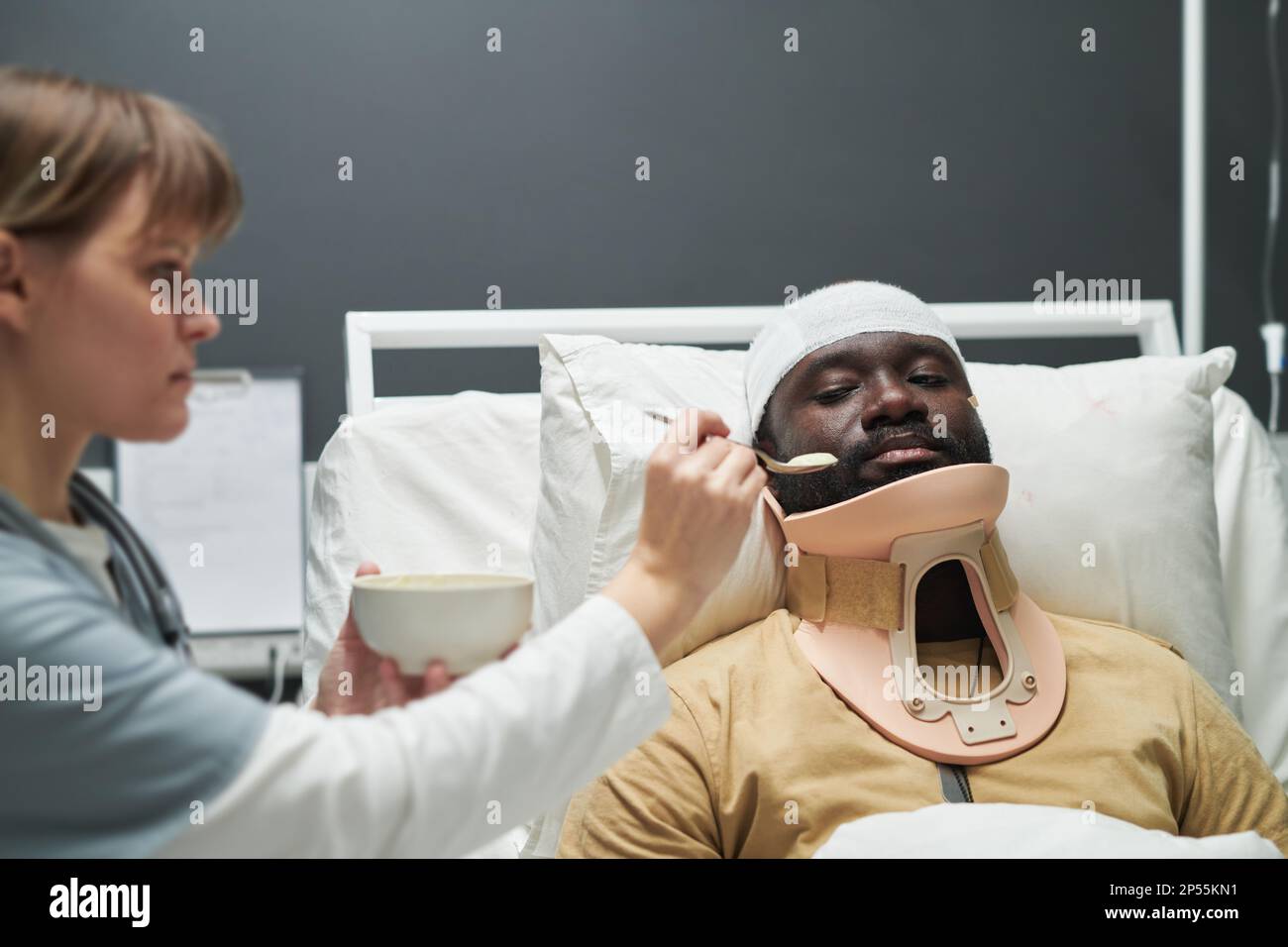 Jeune infirmière tenant une cuillère avec de la nourriture par la bouche d'un patient mâle blessé avec un collier médical allongé dans le lit à l'hôpital militaire Banque D'Images