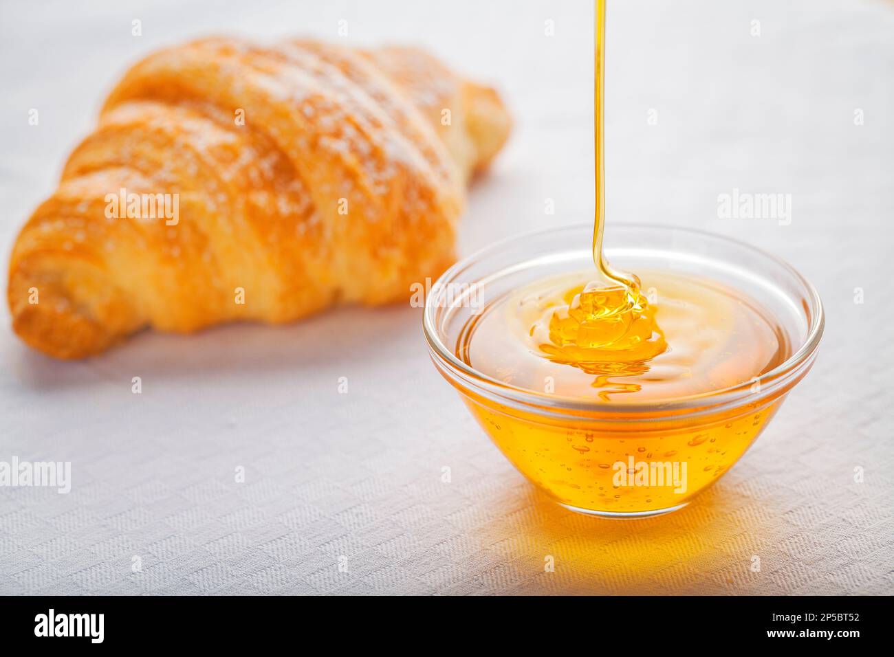 Le miel liquide est versé dans un pot en verre et un croissant flou en arrière-plan. Banque D'Images