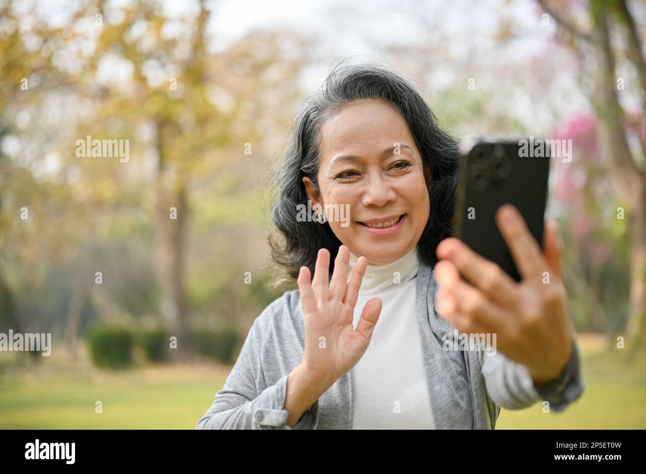 Une femme asiatique heureuse et souriante de 60 ans passe des appels vidéo avec son petit-enfant tout en profitant d'une promenade dans le parc verdoyant. Concept de loisirs et de loisirs Banque D'Images