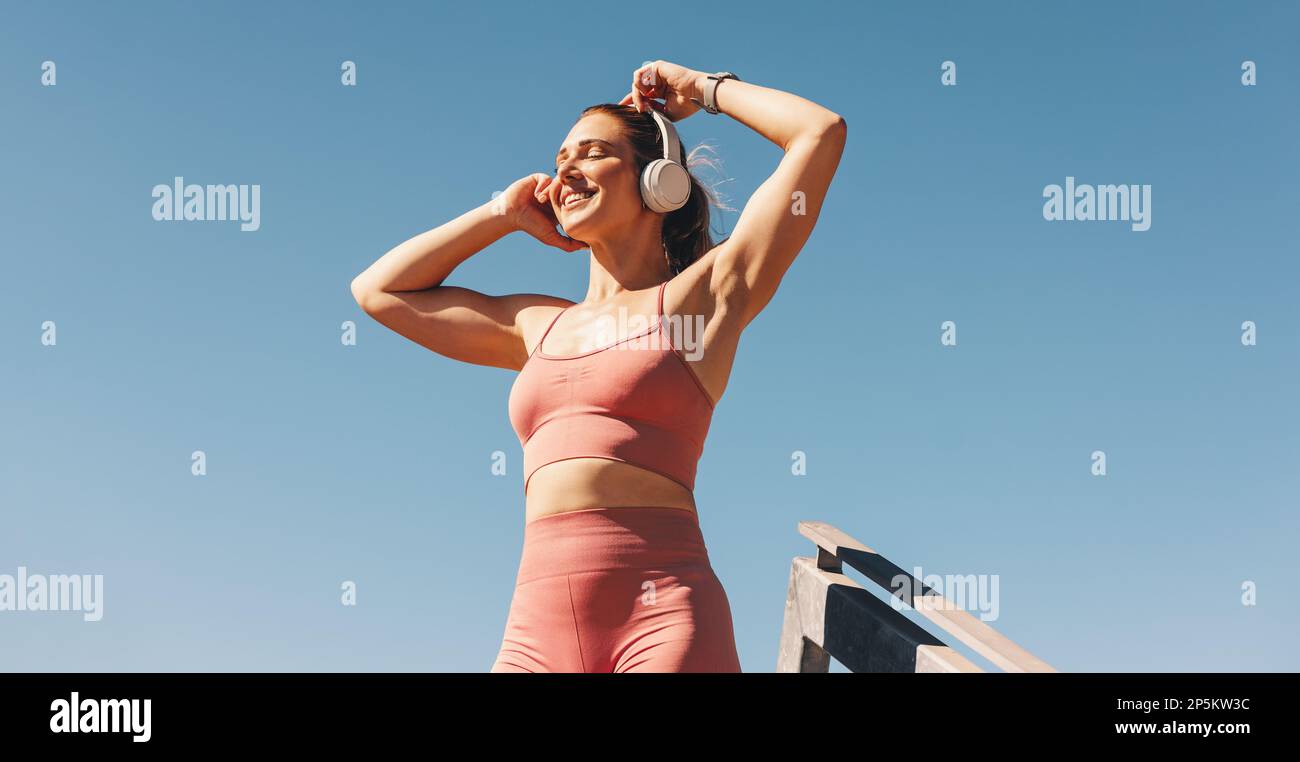 Femme de sport caucasienne écoutant de la musique sur des écouteurs en extérieur. Femme en vêtements de sport debout contre le ciel. Bonne athlète féminine prenant une pause fr Banque D'Images