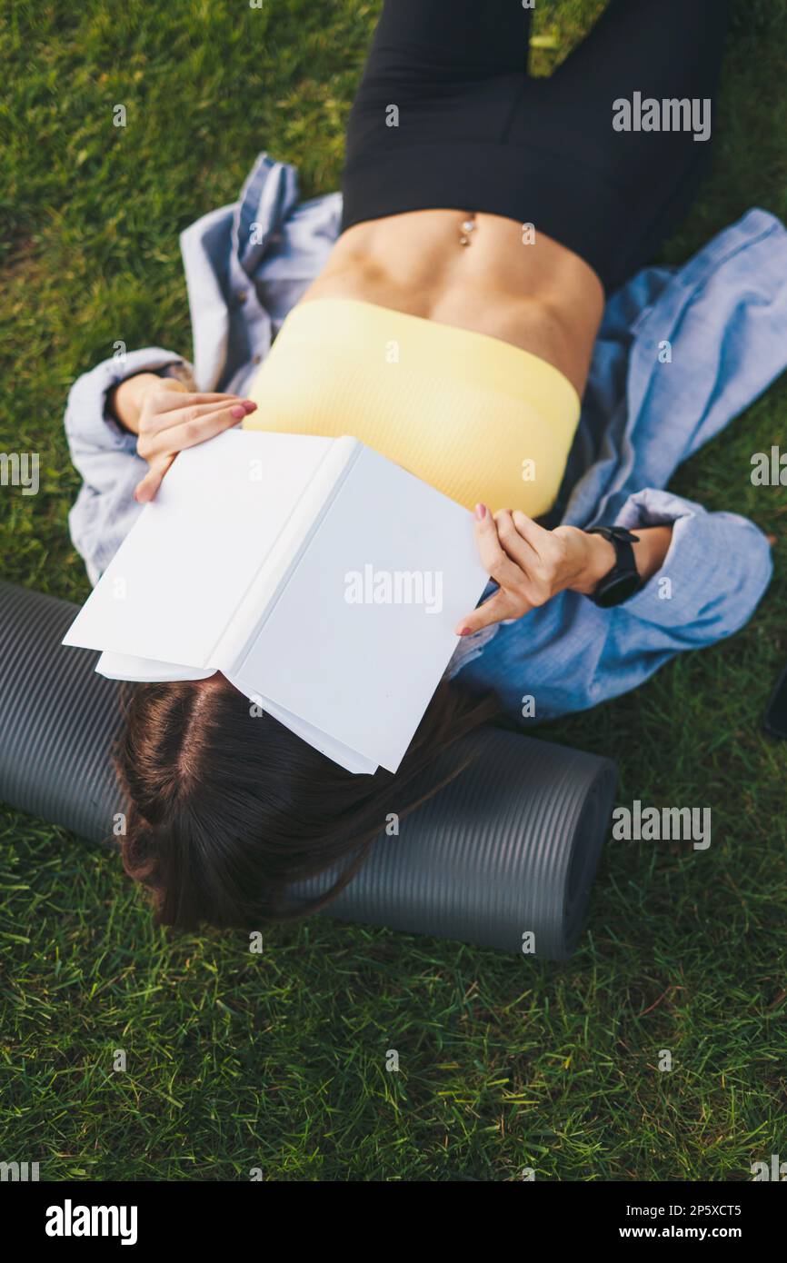 Femme sportive couvrant son visage avec un livre qu'elle lit allongé dans le parc d'été après l'entraînement du matin. Un style de vie heureux. Un mode de vie sain Banque D'Images
