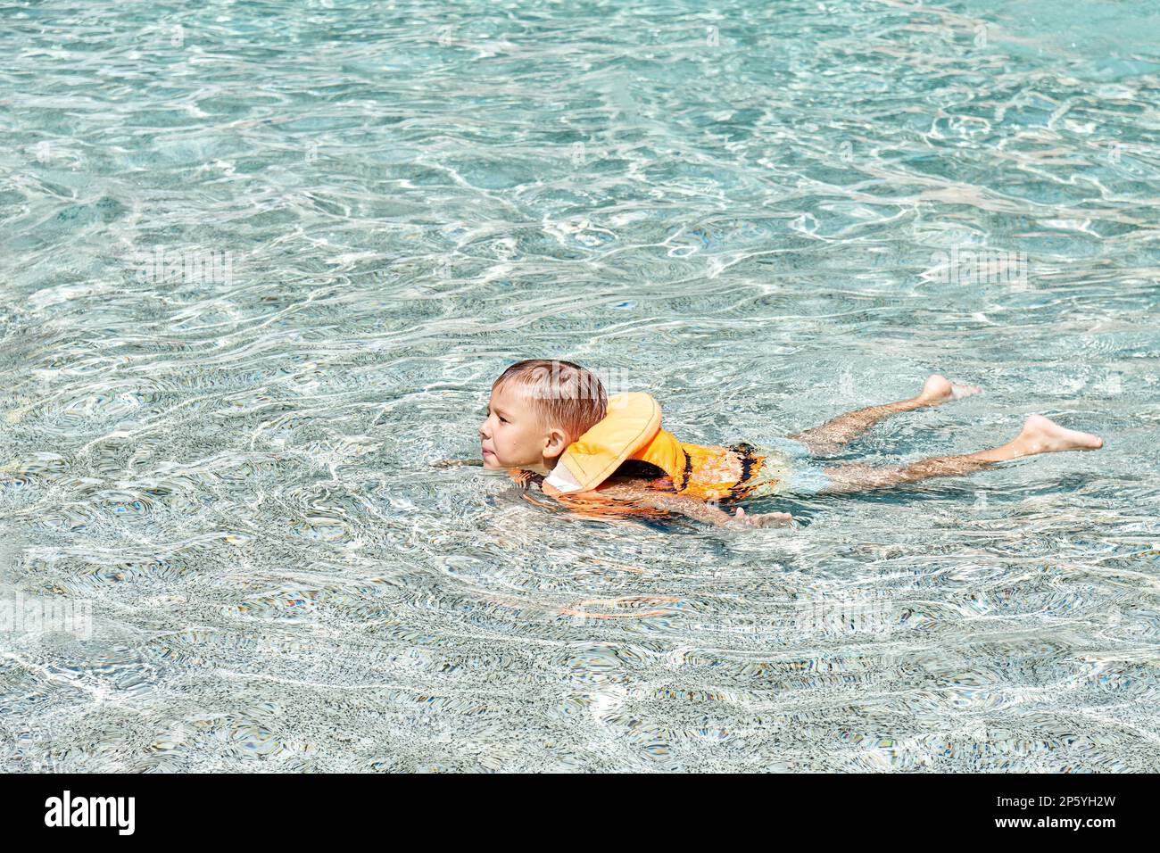 Décontracté, le gilet de sauvetage apprend à nager dans une piscine peu profonde avec de l'eau bleue claire. Garçon aime l'activité dans le parc aquatique pendant les vacances d'été Banque D'Images