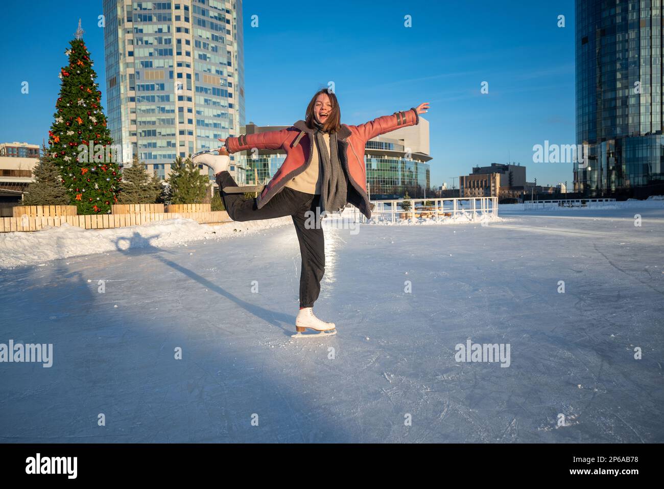 une jeune belle fille sur skate dans un manteau de peau de mouton sur une patinoire avec la toile de fond de la métropole et les rayons du soleil. Banque D'Images