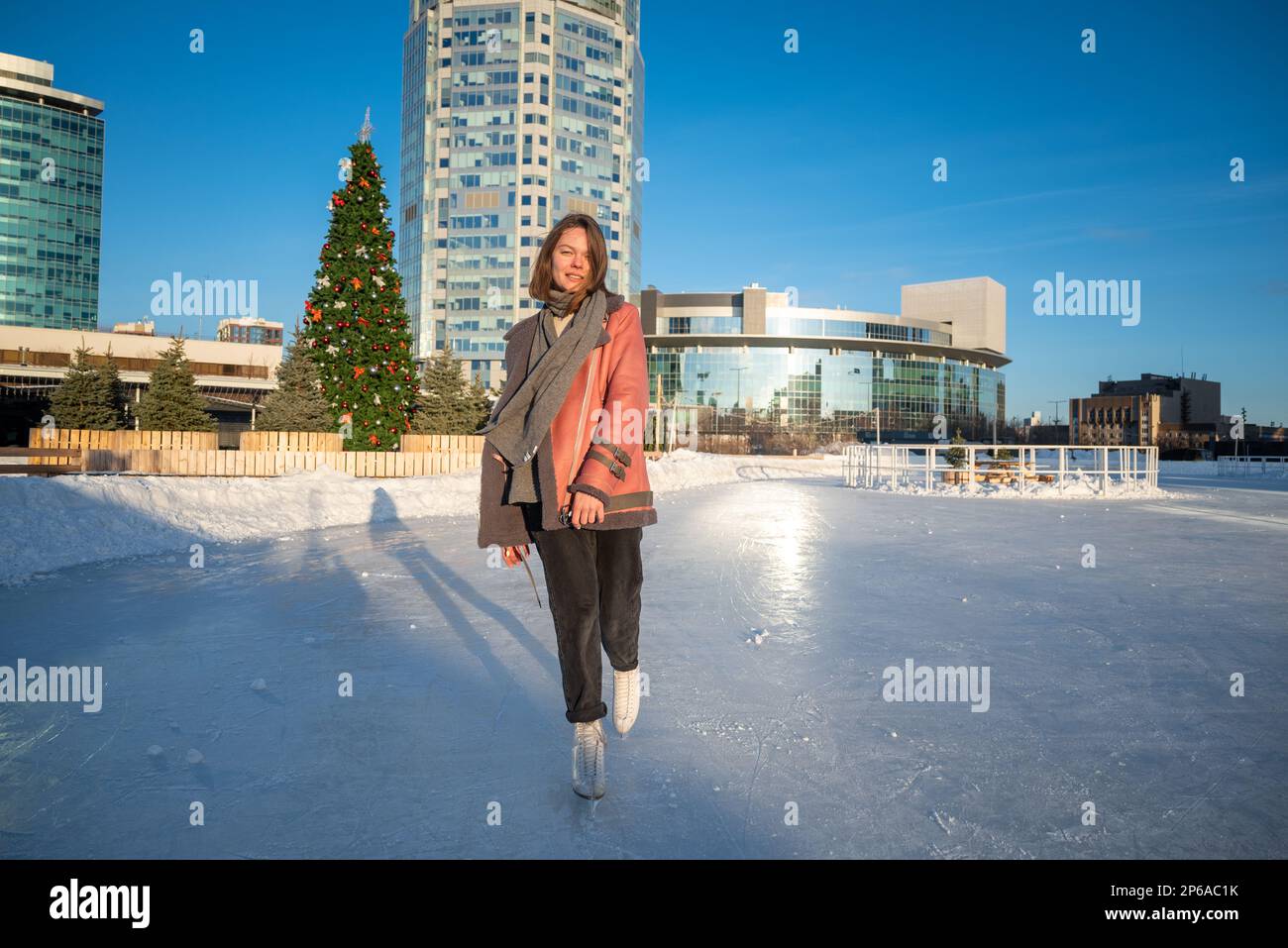 une jeune belle fille sur skate dans un manteau de peau de mouton sur une patinoire avec la toile de fond de la métropole et les rayons du soleil. Banque D'Images