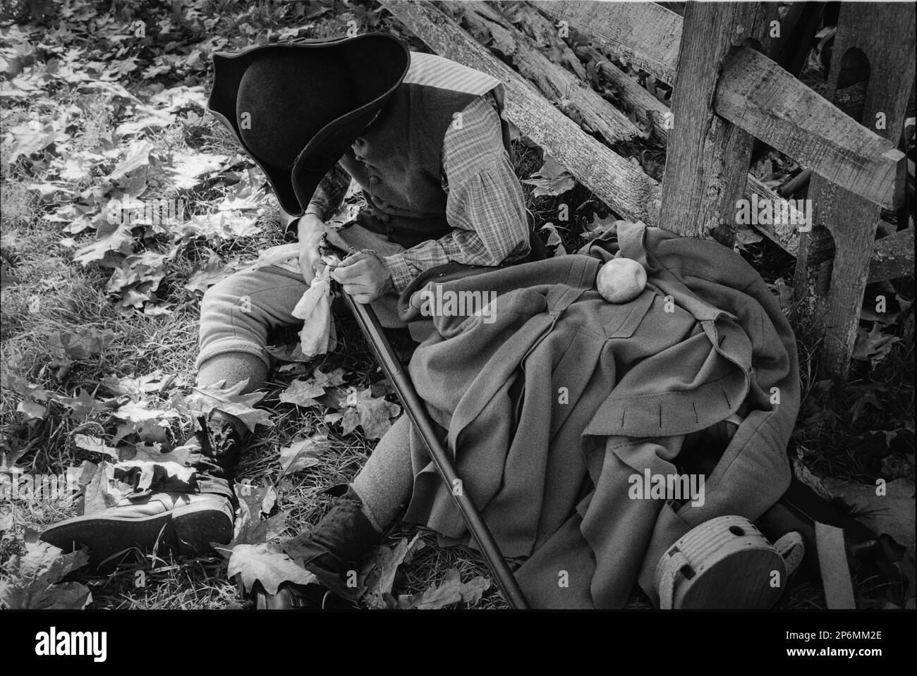 Un acteur en vêtements d'époque et un chapeau de tricorne nettoie son musket tout en se penchant contre une clôture de poteau de bois pendant une reconstitution de la guerre d'indépendance à la Banque D'Images