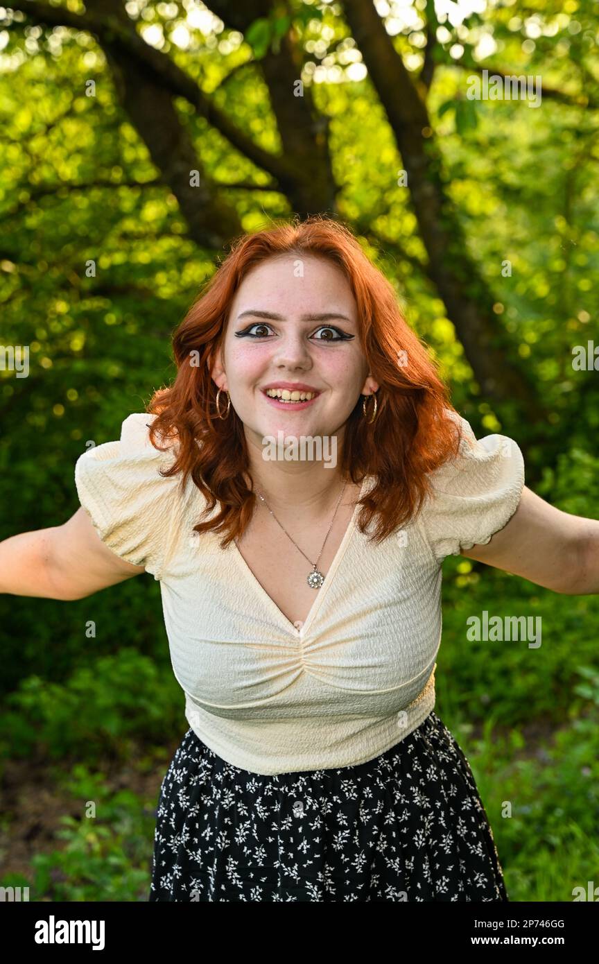 Une jeune femme riant à la chevelure rouge, portant un chemisier de couleur claire, bras étirés sur le côté, dans la nature verte Banque D'Images