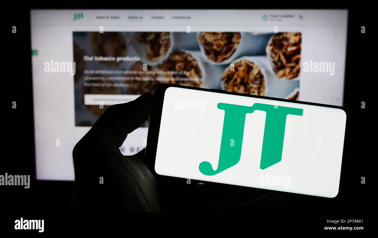 Personne tenant un téléphone portable avec le logo de la compagnie de cigarettes Japan Tobacco Inc (JT) à l'écran en face de la page Web de l'entreprise. Mise au point sur l'affichage du téléphone. Banque D'Images