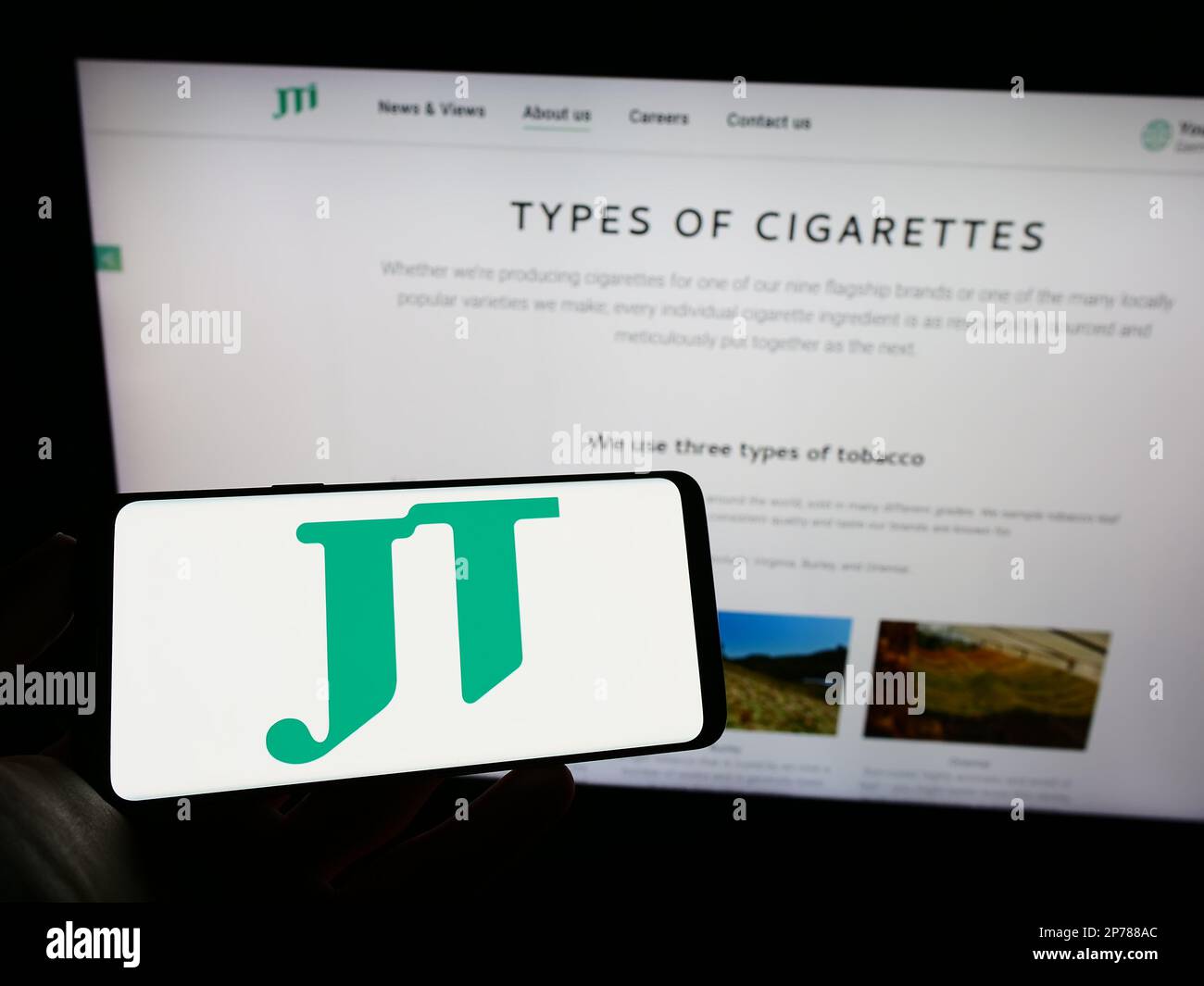 Personne tenant un téléphone cellulaire avec le logo de la compagnie de cigarettes Japan Tobacco Inc (JT) à l'écran en face de la page Web d'entreprise. Mise au point sur l'affichage du téléphone. Banque D'Images