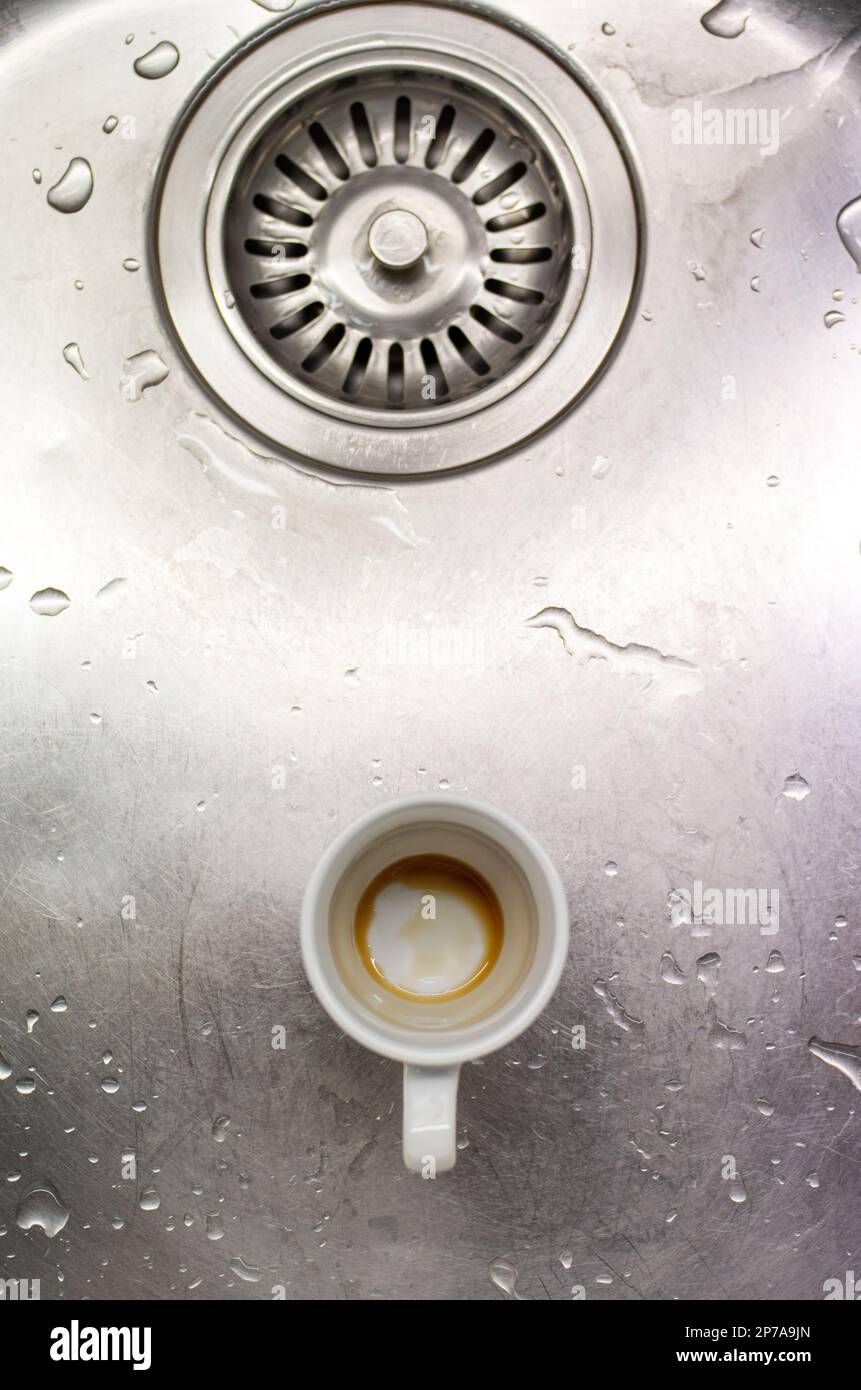 Petite tasse de café sale en porcelaine blanche dans un évier de cuisine en métal à côté du trou d'évacuation vue du dessus. Banque D'Images