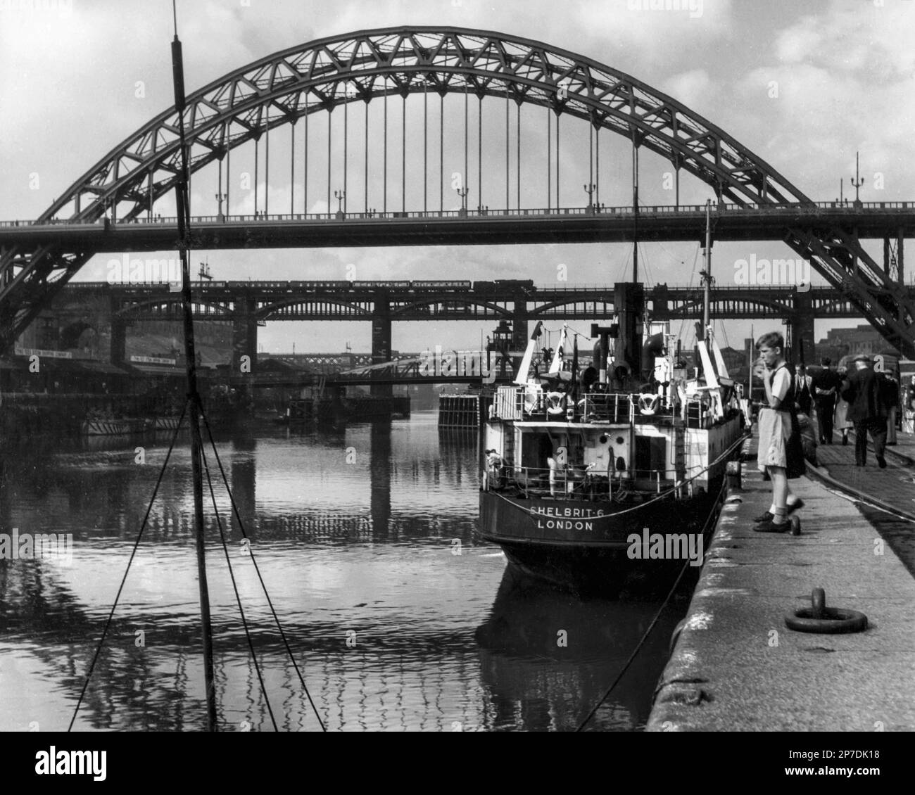 Steam Age Newcastle upon Tyne, Tyne and Wear, Angleterre, Royaume-Uni : une locomotive de débourrage tire des voitures sur High Level Bridge au-delà du pont voûté Tyne et du Quayside de la ville, où Shelbrit 6, un pétrolier côtier Shell-Mex et BP, est amarré. Construit en 1944, le navire à vapeur de 797 tonnes transportait de l'huile pour Shell 1947-1964 et fut mis au rebut en 1973 à Palerme, en Italie. Photographie monochrome vintage 1950. Banque D'Images