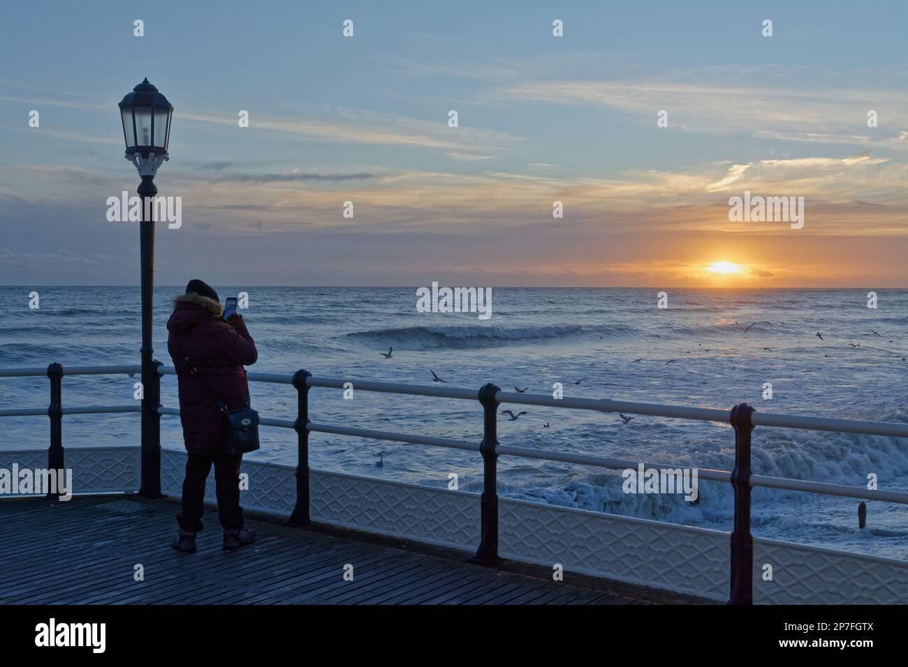 Une femme photographiant un coucher de soleil spectaculaire avec un téléphone, vue depuis Worthing Pier, West Sussex, Angleterre, Royaume-Uni Banque D'Images