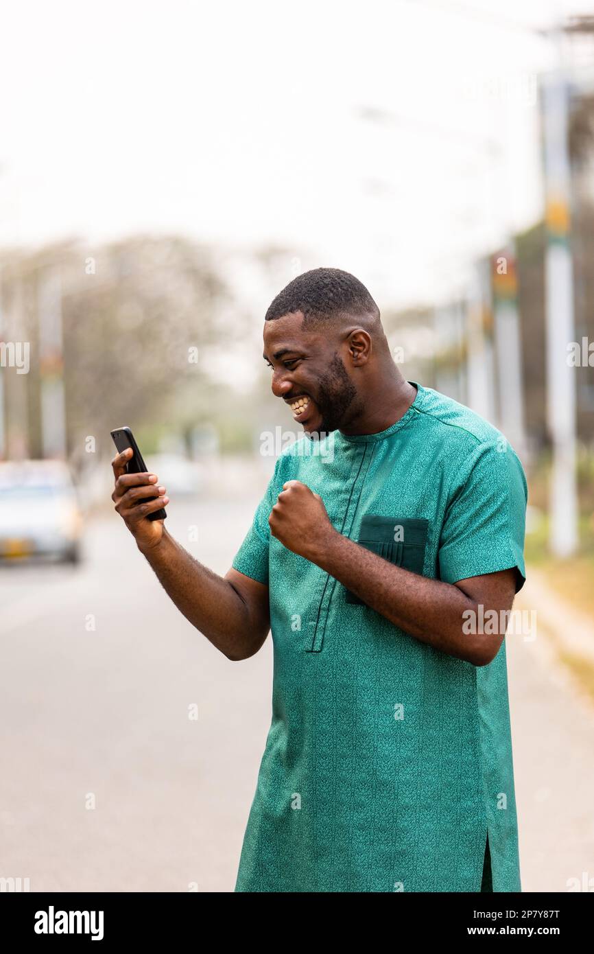 Happy Young African Man Checks Examens scolaires Résultats en ligne avec smartphone, enthousiaste à l'idée de gagner l'admission à l'université pour l'enseignement de troisième cycle. Banque D'Images