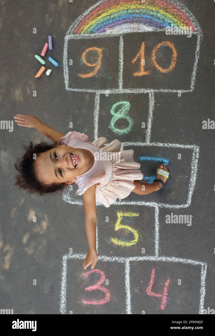 Petite fille afro-américaine et hopscotch coloré dessiné avec de la craie sur l'asphalte à l'extérieur, vue de dessus. Bonne enfance Banque D'Images