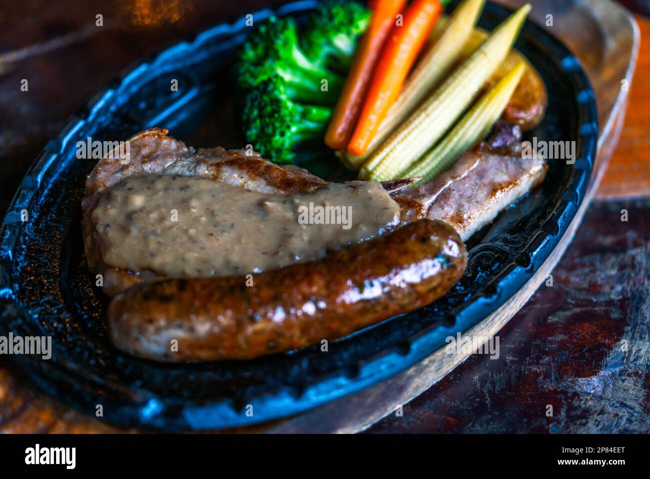 Gros plan sur le steak de porc haché poivre, saucisse, pomme de terre et légumes sur plaque de fer chaude, ancienne table en bois, lumière naturelle. Banque D'Images