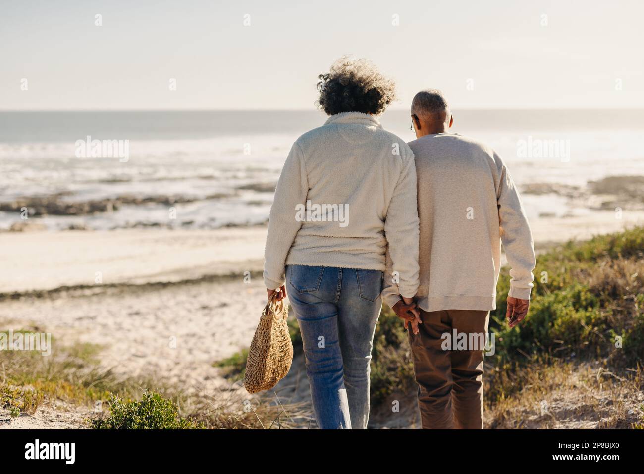 Vue arrière d'un couple romantique senior tenant les mains tout en marchant vers la plage pour un pique-nique. Couple âgé insouciant prenant des vacances au bord de la mer à l'arrivée Banque D'Images