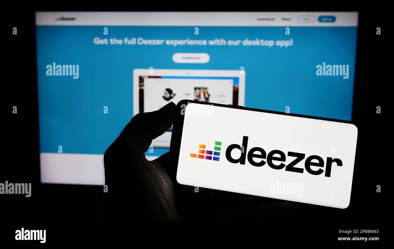 Personne tenant un téléphone portable avec le logo de la société française de streaming musical Deezer S.A. sur écran en face de la page web d'affaires. Mise au point sur l'affichage du téléphone. Banque D'Images