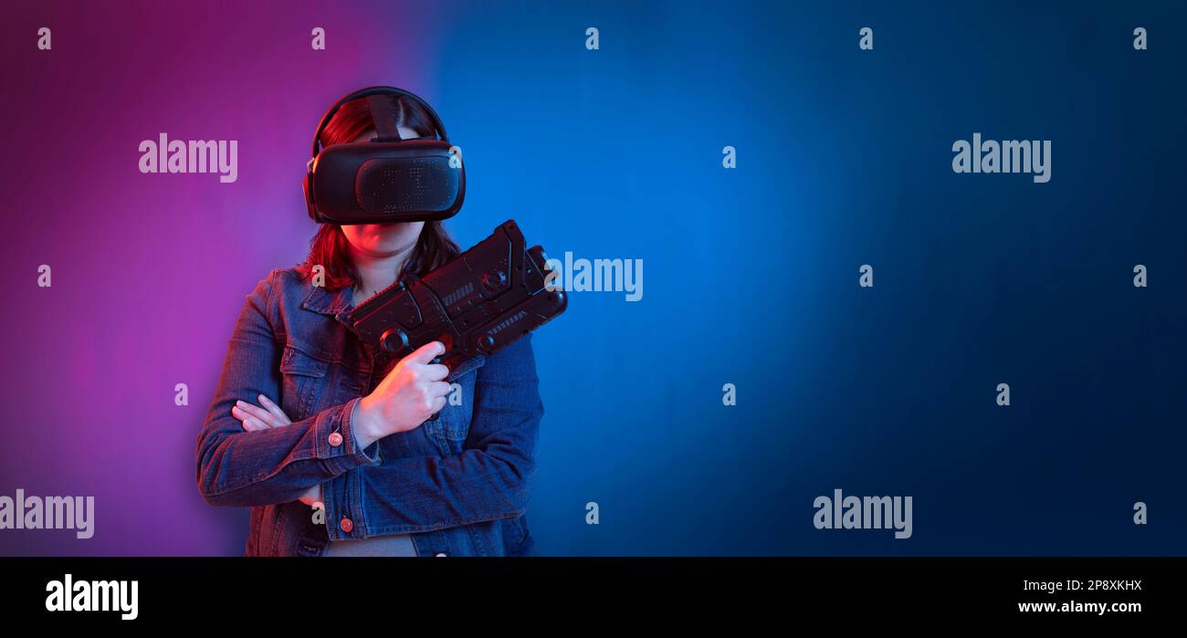 Belle jeune femme hispanique debout vue de face en utilisant des lunettes de réalité virtuelle et un fusil de jeu dans ses mains contre le fond de mur avec bleu et purpl Banque D'Images