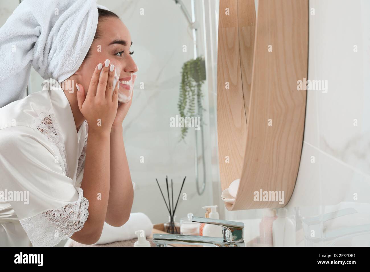 Belle jeune femme appliquant de la mousse nettoyante sur le visage près du miroir dans la salle de bains, espace pour le texte. Cosmétiques pour les soins de la peau Banque D'Images