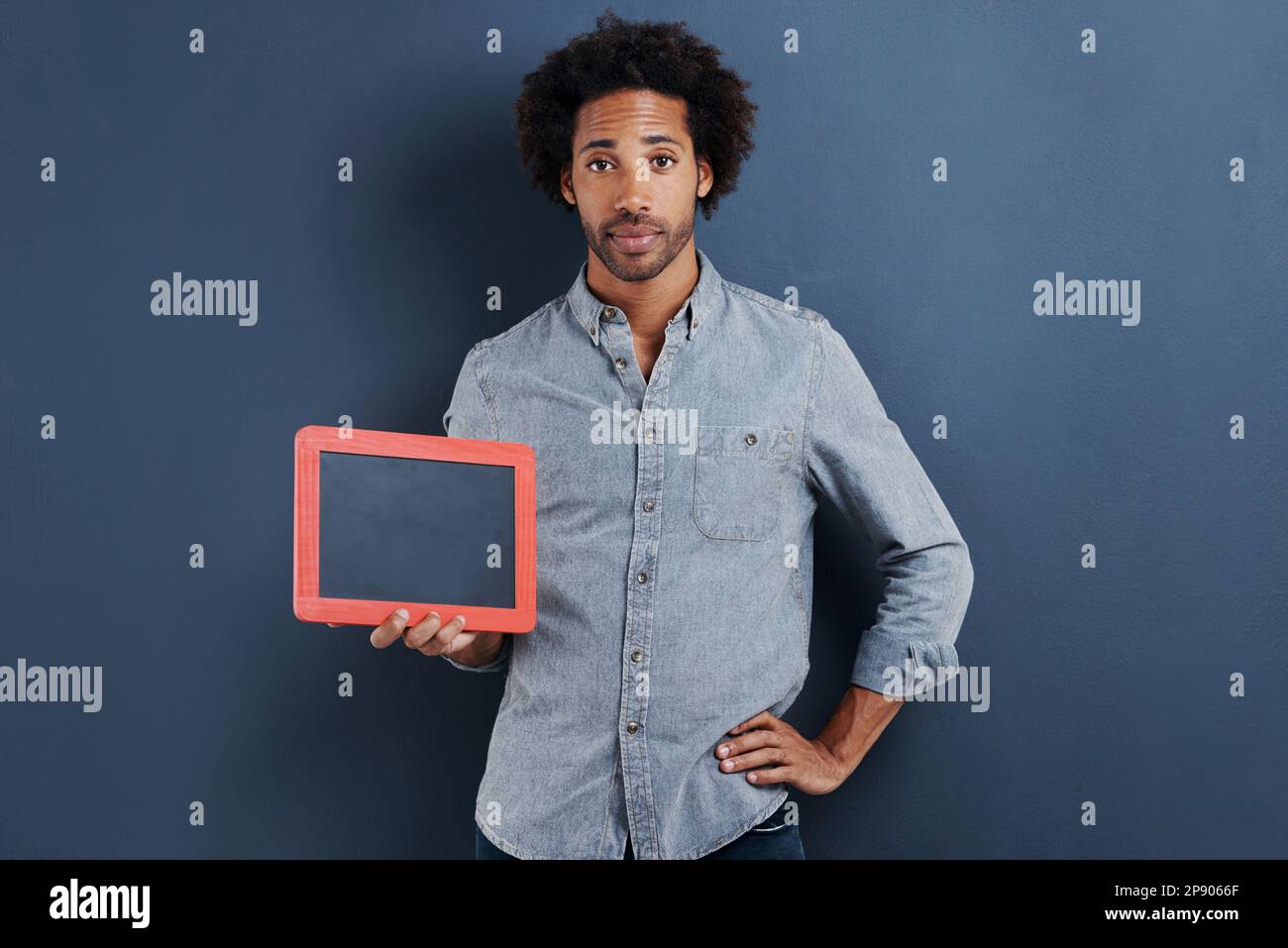 Vous donner un signe. Portrait d'un beau jeune homme avec sa main sur la hanche tenant un petit tableau de craie sur fond gris. Banque D'Images