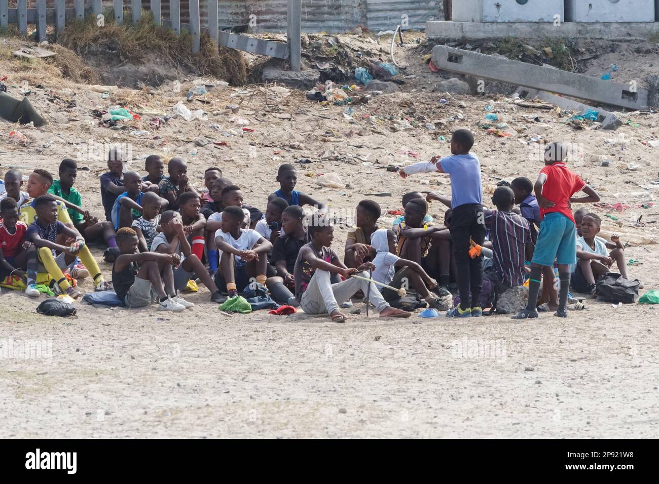 Un groupe de jeunes enfants ou enfants noirs d'Afrique du Sud s'assoient sur le sable pollué et jonché dans une commune en attendant de jouer à un match sportif Banque D'Images