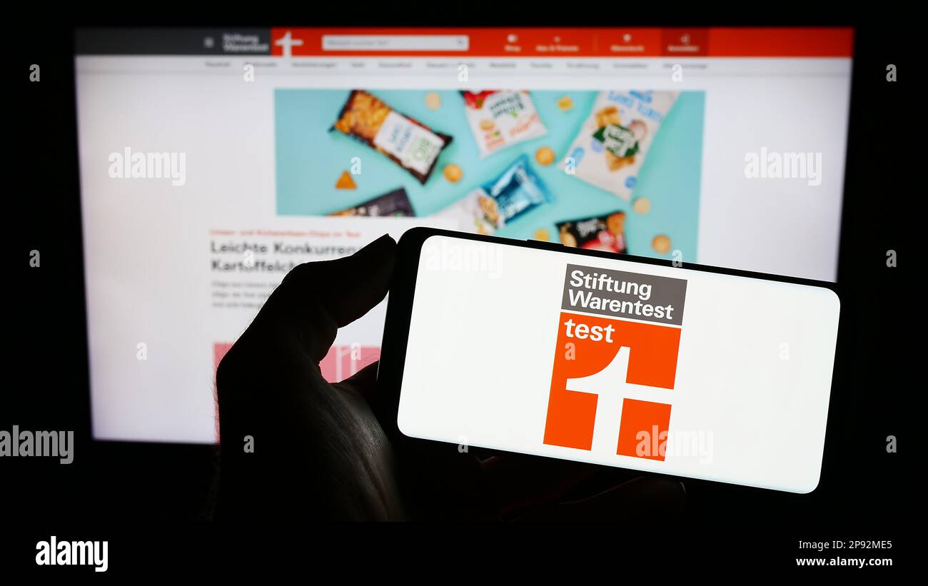 Personne tenant un téléphone portable avec le logo de l'organisation allemande des consommateurs Stiftung Warentest à l'écran en face de la page web. Mise au point sur l'affichage du téléphone. Banque D'Images