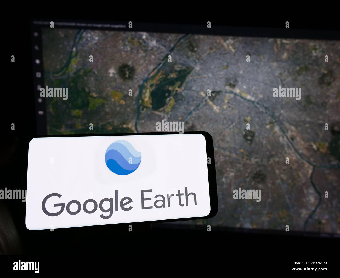 Personne tenant le téléphone portable avec logo du logiciel d'imagerie satellite Google Earth à l'écran en face de la page Web. Mise au point sur l'affichage du téléphone. Banque D'Images