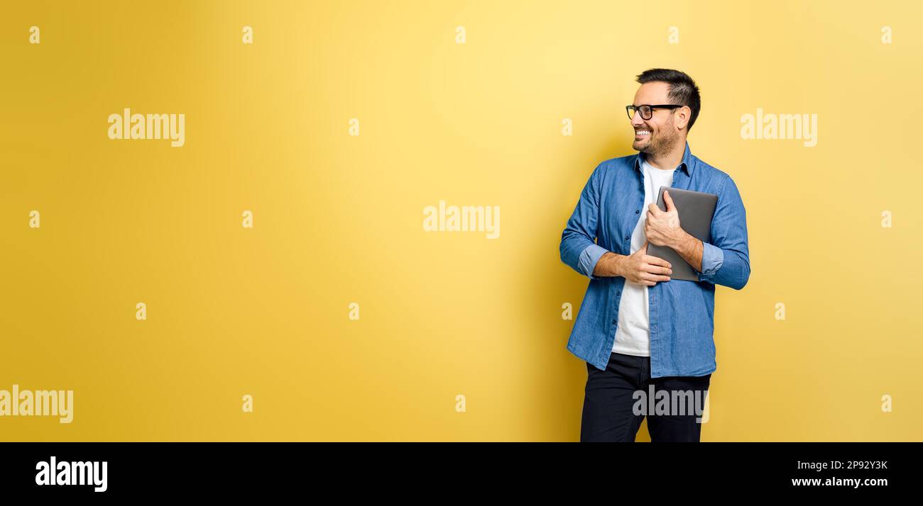 Homme souriant tenant un ordinateur portable et regardant loin. Indépendant positif debout sur fond jaune. Il porte du denim et des lunettes bleus. Banque D'Images