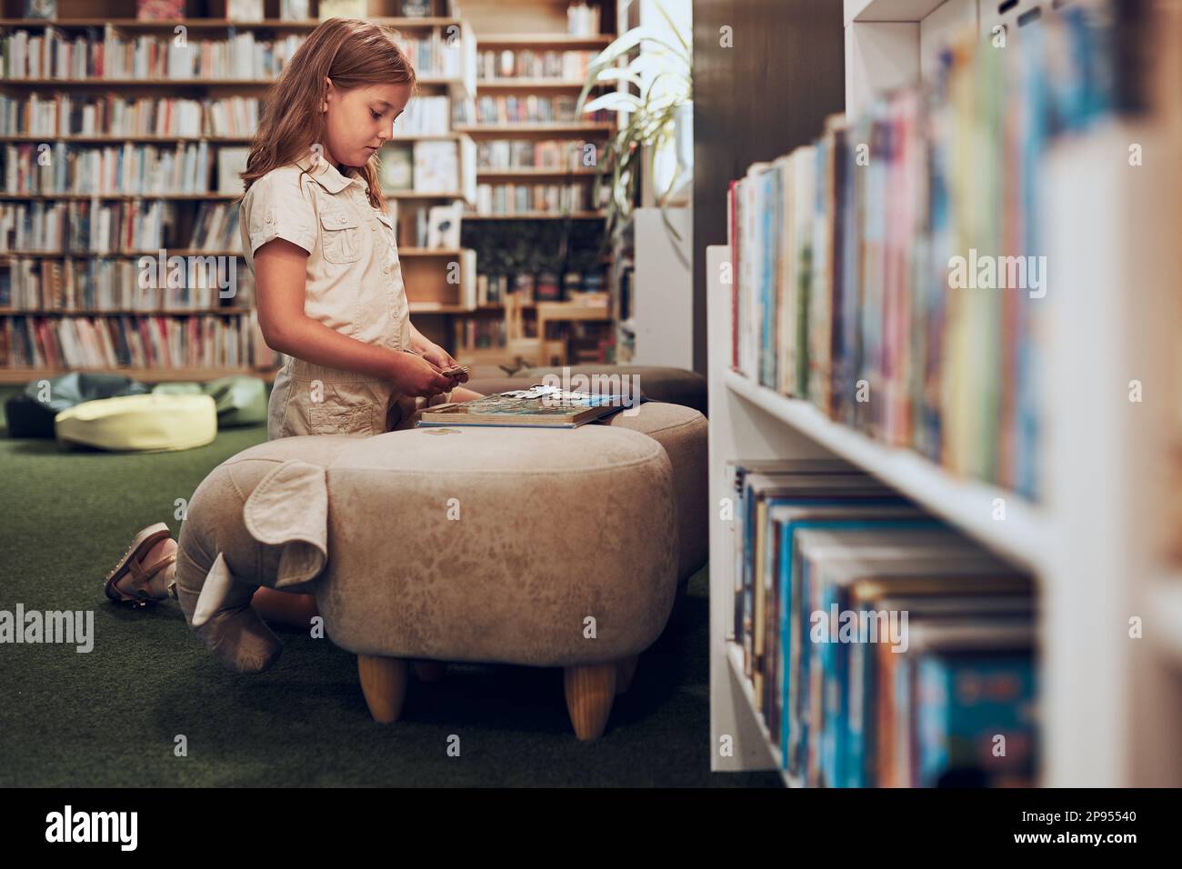 Écolière qui fait du puzzle et livre de lecture dans la bibliothèque de l'école. L'élève de l'école primaire est impliqué dans le livre avec puzzle. Smart girl apprendre à partir du livre. Benef Banque D'Images