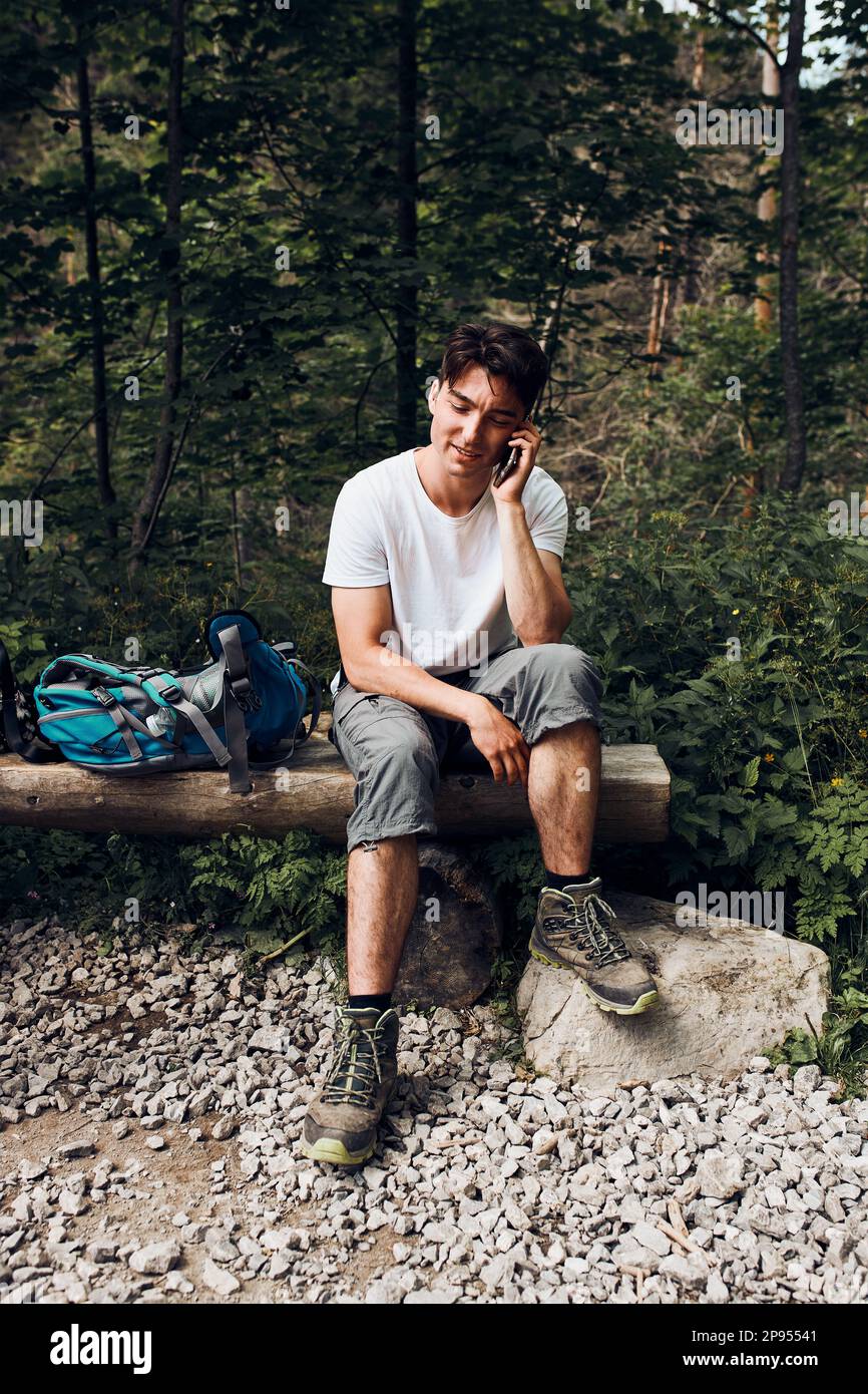 Jeune homme avec sac à dos parlant au téléphone ayant une pause assis sur le coffre pendant la randonnée dans une montagne qui passe activement des vacances d'été Banque D'Images
