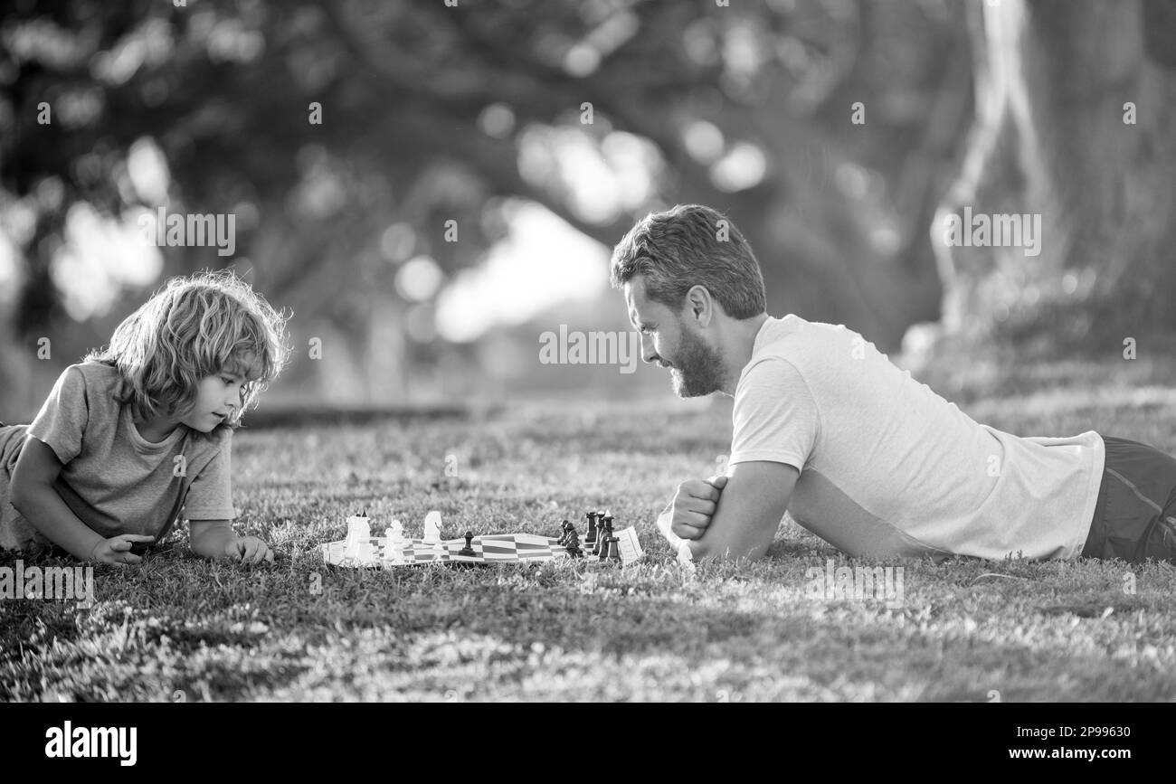 bonne famille de papa et garçon de fils jouant aux échecs sur l'herbe verte dans le parc à l'extérieur, concentration. Banque D'Images