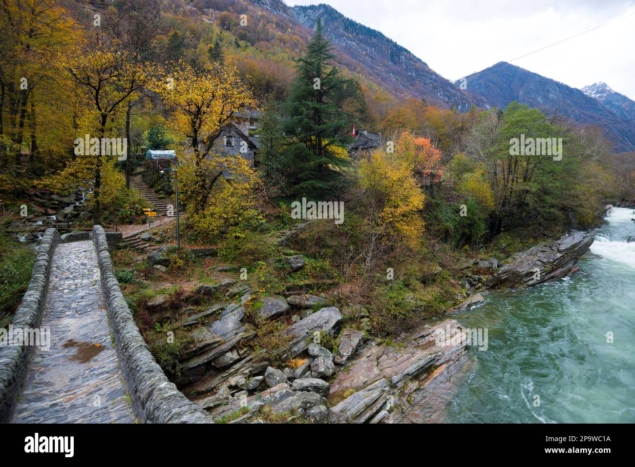 Pont romain sur la rivière qui coule rapidement dans le Val Verzasca, Suisse, jour d'automne nuageux, arbres colorés sur les pentes de montagne et maisons en pierre Banque D'Images