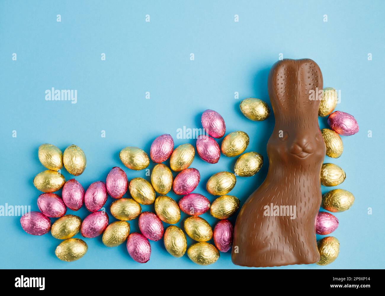 Grand lapin au chocolat au lait et pile d'œufs de Noël en papier aluminium enveloppé de rose et d'or sur fond bleu. Concept joyeuses Pâques. Préparation de l'hologramme Banque D'Images
