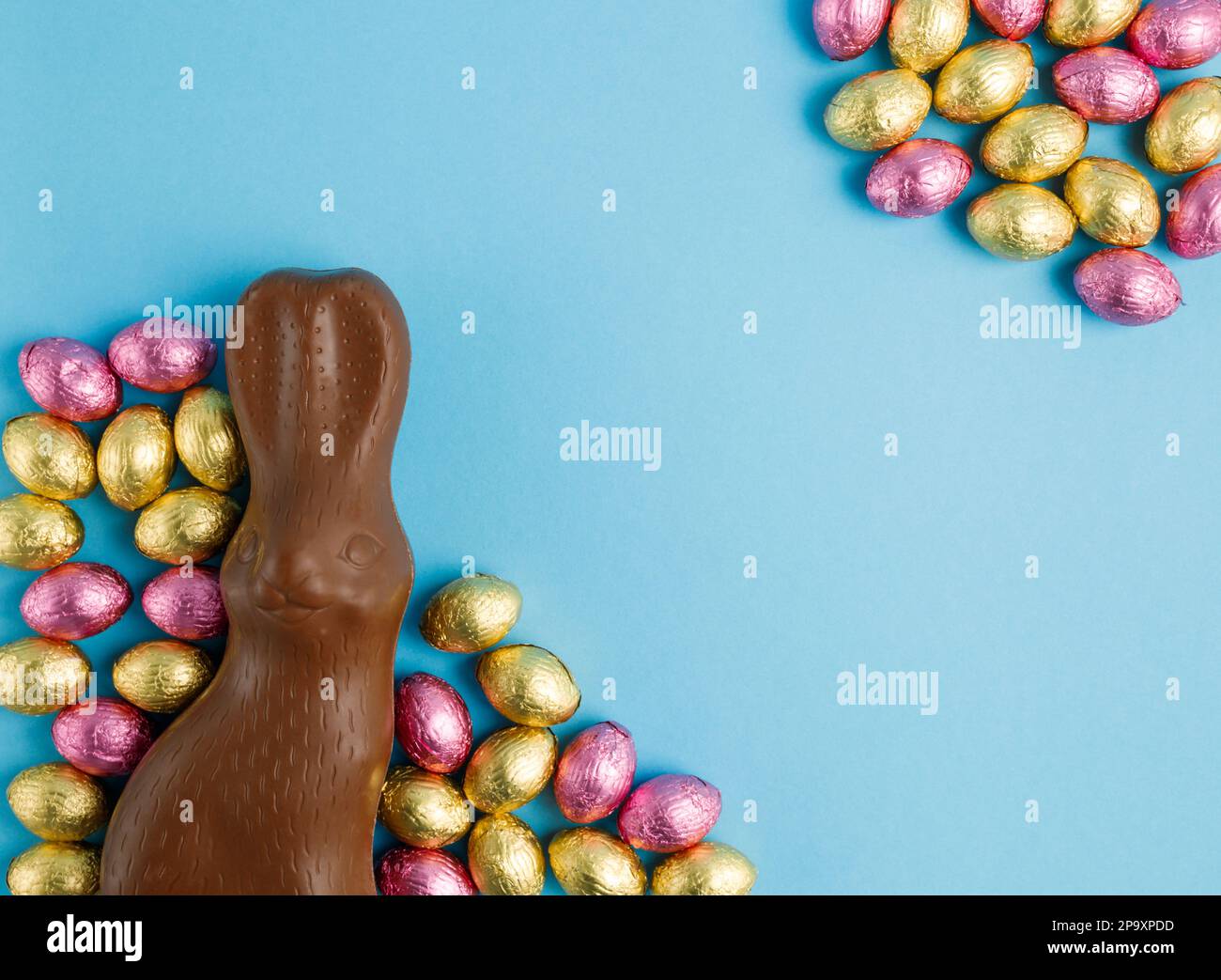 Big Milk chocolat lapin de Pâques et cadre de pile d'œufs de bonbons enveloppés dans du papier d'aluminium rose et doré sur fond bleu. Concept joyeuses Pâques. Préparation Banque D'Images