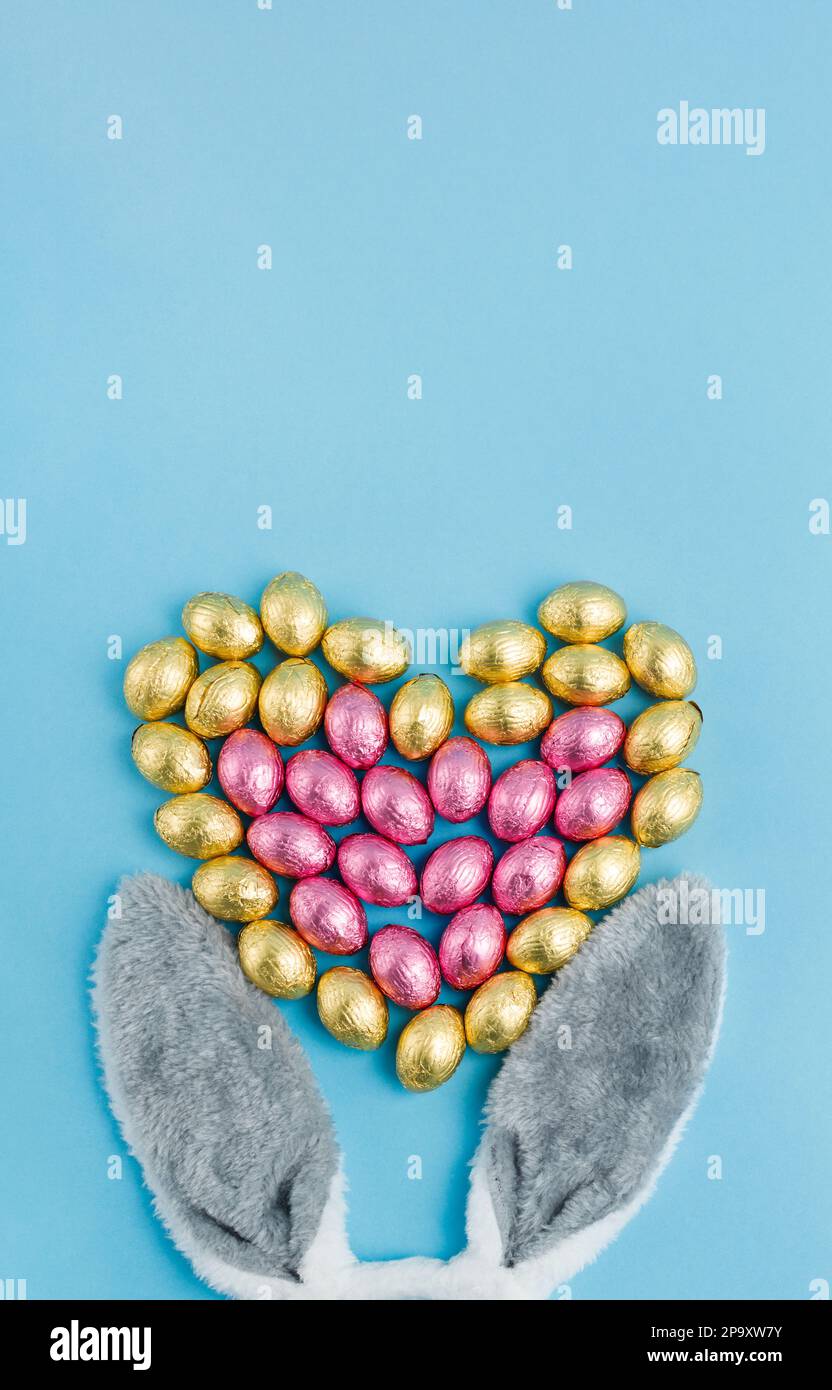 Oreilles de lapin de Pâques et pile d'œufs en forme de cœur enveloppés de papier d'aluminium rose et doré sur fond bleu. Joyeuses Pâques, concept amour. Préparation pour Banque D'Images