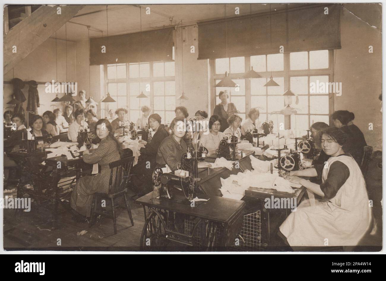 Photo du début du 20th siècle d'un groupe de travailleurs du textile. Les femmes sont assises aux machines à coudre à pédales dans un atelier avec une grande fenêtre Banque D'Images