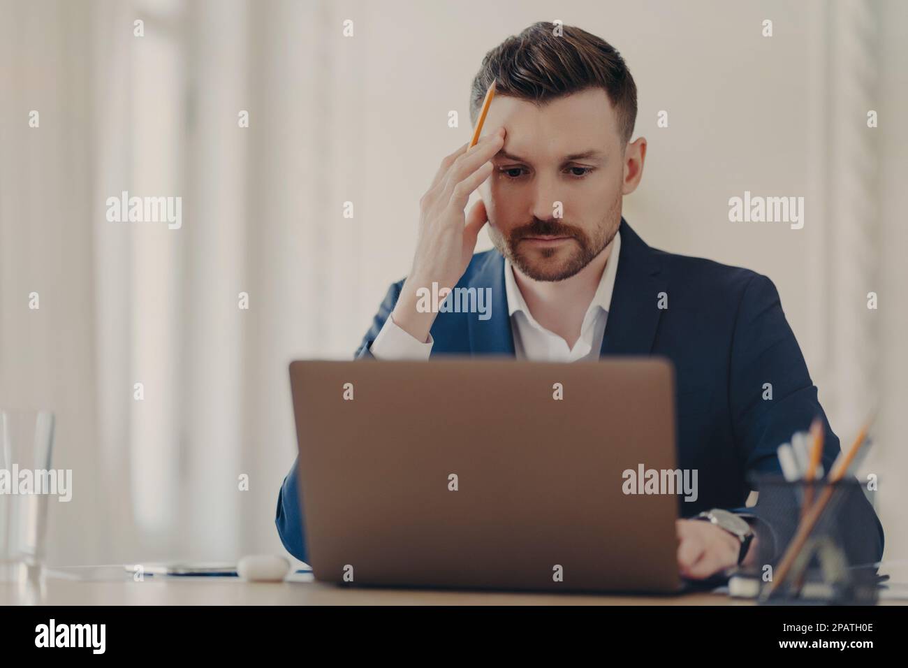 Beau homme d'affaires réussi et réfléchi en costume bleu foncé assis à son bureau dans un bureau de couleur claire travaillant sur le plan d'affaires, tenant un crayon Banque D'Images